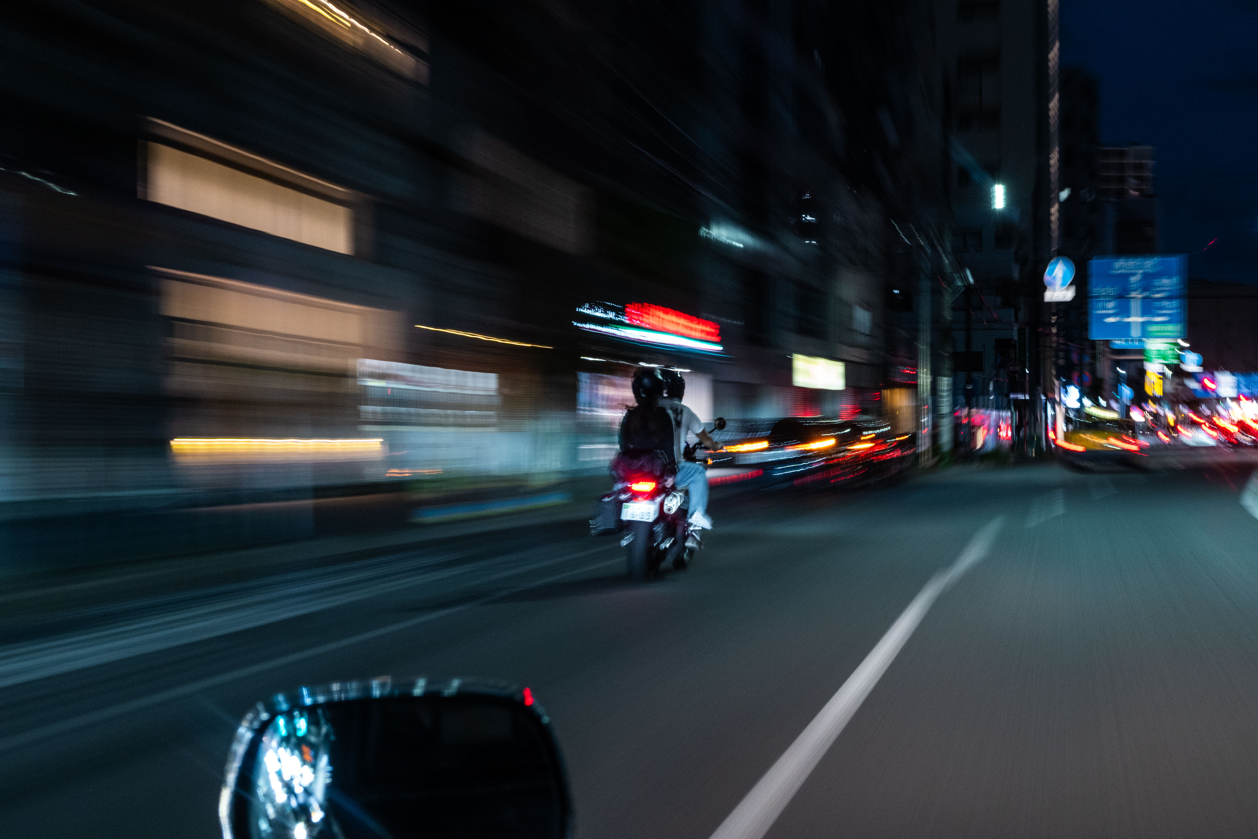 A motion-blurred night photograph from inside a moving vehicle on a Tokyo street. A scooter rider in dark jacket rides ahead, slightly in focus. Around them the city smears into streaks of electric blue, red and orange — neon signs and shopfronts and tail-lights drawn out by speed. A wing mirror at the bottom edge of the frame.