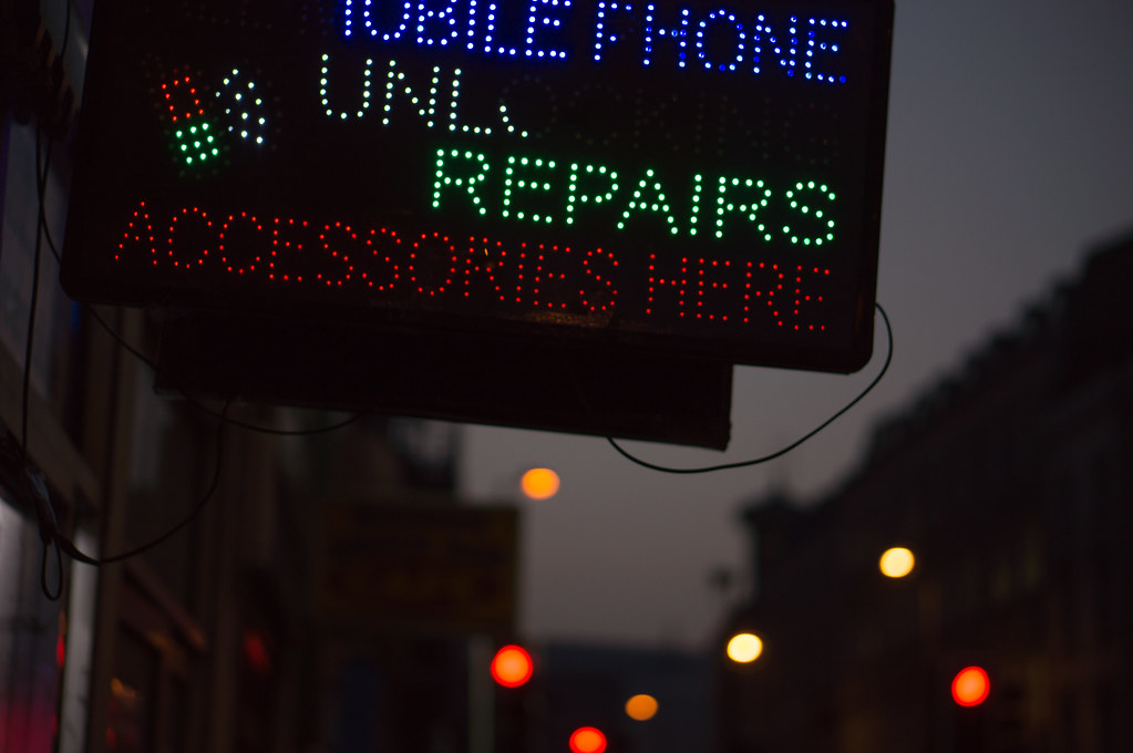 A photograph at dusk of an LED dot-matrix shop sign reading MOBILE PHONE UNLOCKING REPAIRS ACCESSORIES HERE in flickering blue, green and red dots. Behind it, soft orange and red bokeh of street lights along a row of buildings.