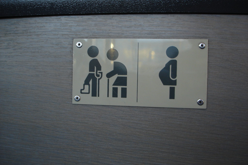 A polished metal sign with three black pictograms on a brushed cream ground: a standing figure with a cane, a seated figure leaning forward, and a pregnant woman in profile. Mounted to a wood-grain wall panel by four silver studs.