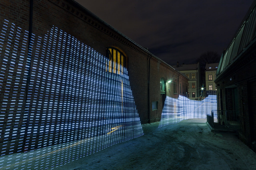 A long-exposure photograph at night: a brick courtyard between buildings in Oslo, snowy ground, a yellow-lit window. Filling the frame are two enormous walls of vertical blue-white light bands, the WiFi field in the courtyard rendered as a 3D topography by the four-metre LED rod walked through it.