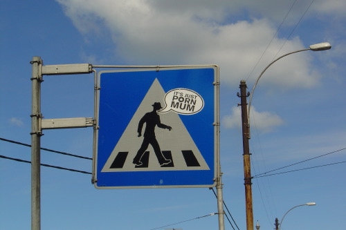 A blue Norwegian pedestrian-crossing sign in Majorstua, Oslo, the white-on-blue figure mid-stride across the white triangle. A small black-ink speech bubble has been stickered onto the sign at head height: 'IT'S JUST PORN MUM'. Sky, a streetlight, overhead wires.