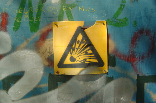 A yellow industrial-warning sign (black triangle, exploding-bomb pictogram inside) bolted to a wall and surrounded by graffiti: green spray paint above reading 'HEGRER OG EN MUS' (Norwegian for 'herons and a mouse'), white silver tags overlaid on a turquoise-and-red wall. The official sign sits in the middle of the unauthorised marks, both speaking the same language of the surface.